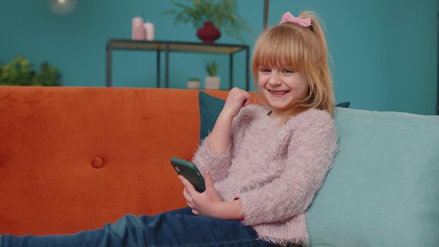 Happy one little school kid girl use mobile cell phone browsing say wow yes found out great big win news. Excited child doing winner gesture celebrate holding smartphone lying on sofa alone at home