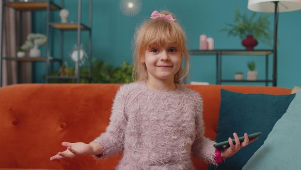 Excited amazed kid girl use mobile cell phone say wow yes raising hands in surprise looking at camera with big eyes, shocked by sudden victory. Excited winner child celebrate on sofa alone at home