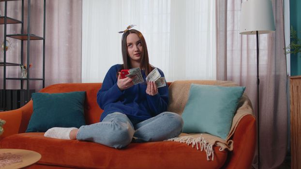 Planning family budget. Smiling happy young woman holding cash money and mobile phone calculate domestic bills at home. Joyful girl satisfied of income and saves money for a planned vacation, gifts