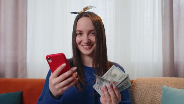 Planning family budget. Smiling happy young woman holding cash money and mobile phone calculate domestic bills at home. Joyful girl satisfied of income and saves money for a planned vacation, gifts