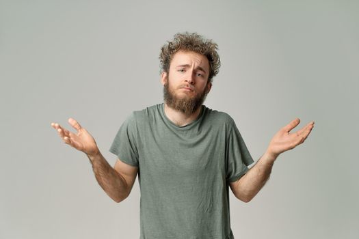 Gesturing I DONT KNOW or I AM SORRY young handsome bearded wild curly hair man with bright blue eyes isolated on grey background. Young thinking man in green t shirt on white. Copy space.