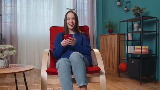 Excited happy joyful stylish teen girl in sweater use mobile cell phone typing browsing say Wow yes found out great big win news doing winner gesture. Young adult woman at home in living room on chair