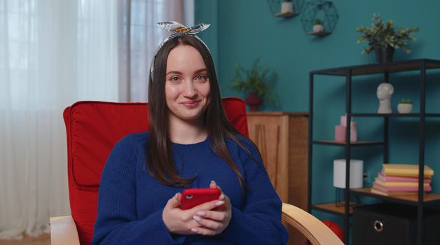 Cheerful relaxing young brunette woman sitting on orange chair uses mobile phone smile at modern home apartment. Happy girl texting share messages content on smartphone social media application online