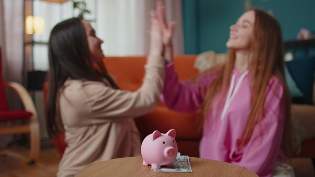 Happy girls friends siblings sitting on floor and take turns dropping dollar banknote into piggy bank, save pocket cash for fun, dreams, future vacation. Keep money, thinking about tomorrow concept