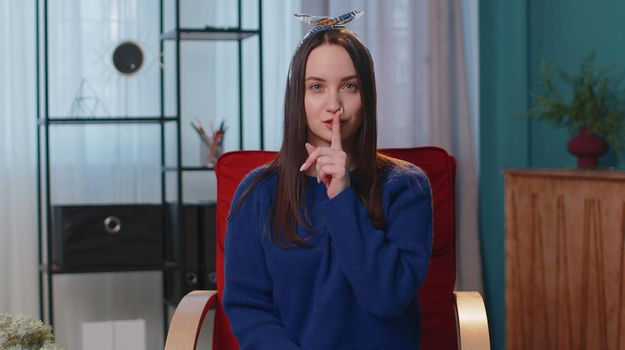 Shh be quiet please. One teenager student girl 20s years old presses index finger to lips makes silence gesture sign do not tells secret. Young adult woman sitting at home in living room on chair