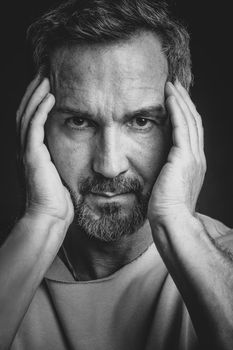 Middle aged grey haired man hold his face with hands looking at camera. Handsome middle aged man in black and white photo. Black and white portrait of attractive, beautiful, serious man.