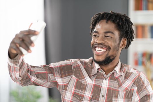 Smiling African American student or freelancer use smartphone having video call or taking selfie. Portrait of happy man with phone in hand looking at smartphone camera. Social media concept.