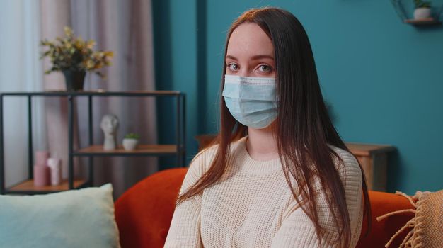 Portrait of young sick girl wearing medical protection mask sitting in living room looking at camera. Depressed female woman spending time alone. Quarantine Coronavirus Pandemic concept. Stay at home