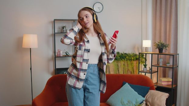 Pretty Caucasian young woman in white sweater in wireless headphones listening music on smartphone, energetic dancing, singing in living room. Indoors. Stay at home. People relax activities concept