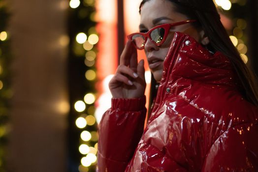 Model fashion girl in sun glasses with braided hair and red glossy coat standing outdoors in night city background. Night life concept. Night club life concept.