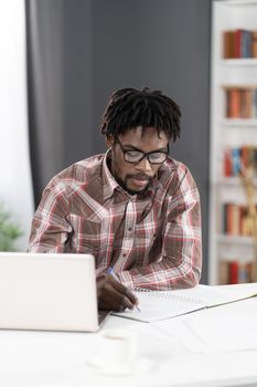 Young african student man making notes working at home working on laptop or listening teacher or boss during video call. African student study using laptop at home.