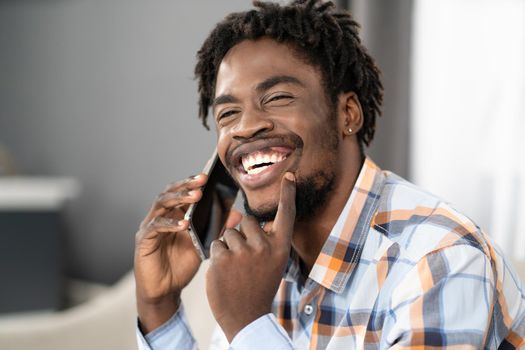 Happy smiling young African American man talking on the phone holding smartphone in hand looking away. Portrait of happy man having present conversation sitting on the sofa. Social media concept.