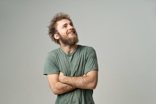 Smiling handsome young man with bearded and wild curly hair, bright blue eyes looking up with hands folded isolated on white background. Young thinking man in green t-shirt on white background.