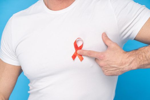 Man hold red ribbon bow AIDS awareness symbol on a heart wearing a white t-shirt isolated on a blue background. Modern medicine and healthcare. AIDS awareness concept.