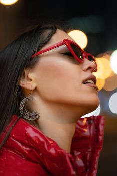 Neon lights night life fashion girl in sun glasses with braided hair and red glossy coat standing outdoors in night city background. Night life concept. Night club life concept.