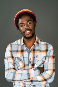 Young smiling black man in afro hat standing with arms folded looking at camera. Cheerful african american young man in plaid shirt looking at camera isolated on gray background.