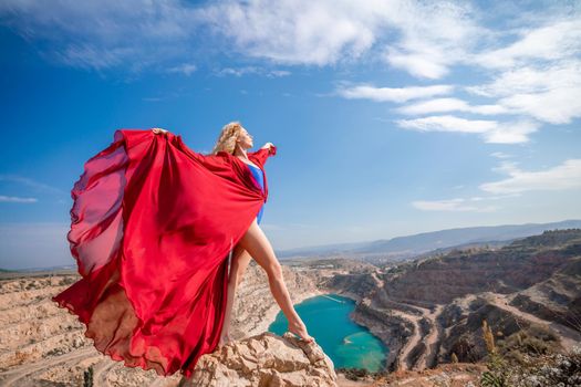 Side view of a beautiful sensual woman in a red long dress posing on a rock high above the lake in the afternoon. Against the background of the blue sky and the lake in the form of a heart.