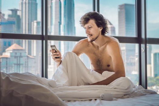 Man wakes up in the morning in an apartment in the downtown area with a view of the skyscrapers and uses smartphone. Life in the noise of the big city concept. Not enough sleep.