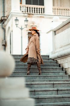 Outdoor fashion portrait of young elegant fashionable brunette woman, model in stylish hat, choker and light raincoat posing at sunset in European city