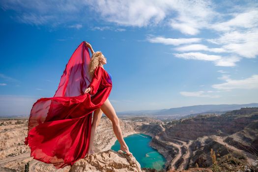 Side view of a beautiful sensual woman in a red long dress posing on a rock high above the lake in the afternoon. Against the background of the blue sky and the lake in the form of a heart.