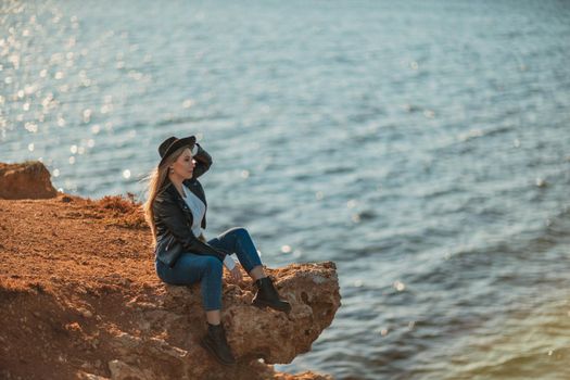 A blonde girl in a stylish black leather jacket is sitting on the seashore