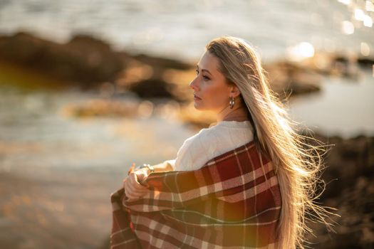 Attractive blonde Caucasian woman enjoying time on the beach at sunset, sitting in a blanket and looking to the side, with the sunset sky and sea in the background. Beach vacation