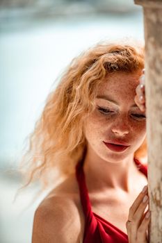Outdoor portrait of a young beautiful natural redhead girl with freckles, long curly hair, in a red dress, posing against the background of the sea
