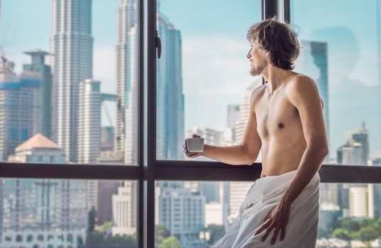 Man wakes up in the morning in an apartment in the downtown area with a view of the skyscrapers and drinks coffee. Life in the noise of the big city concept. Not enough sleep.