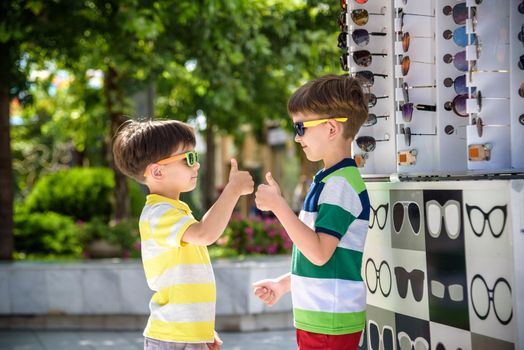 A child and a choice of sunglasses. Two little boys are standing in sun-proof glasses against the background of a shop window with glasses. Kid help choose each other. Sunglasses sale during summer vocation.