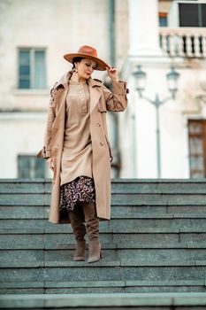 Outdoor fashion portrait of young elegant fashionable brunette woman, model in stylish hat, choker and light raincoat posing at sunset in European city