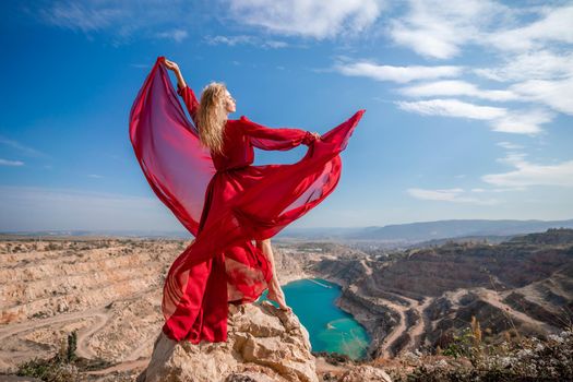 Side view of a beautiful sensual woman in a red long dress posing on a rock high above the lake in the afternoon. Against the background of the blue sky and the lake in the form of a heart.