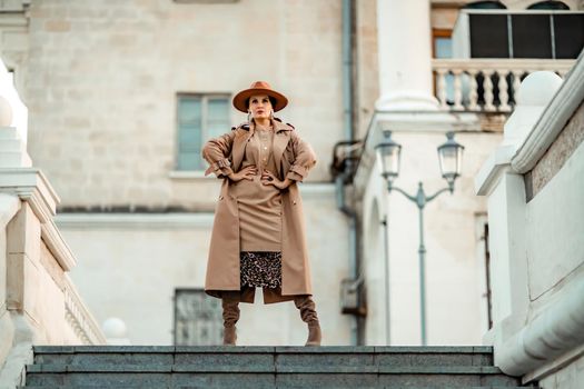 Outdoor fashion portrait of young elegant fashionable brunette woman, model in stylish hat, choker and light raincoat posing at sunset in European city