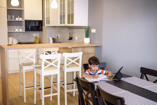 Smart preteen schoolboy doing his homework with digital tablet at home. Child using gadgets on his kitchen to study. Modern education and learning for kids.