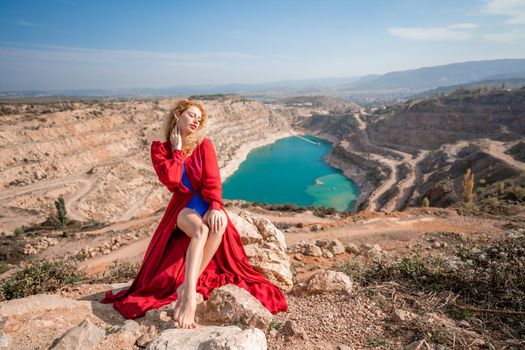 A beautiful girl in a red long dress, Sits on a rock high above the lake in the afternoon. Against the background of the blue sky and the lake in the shape of a heart