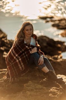 Attractive blonde Caucasian woman enjoying time on the beach at sunset, sitting in a blanket and looking to the side, with the sunset sky and sea in the background. Beach vacation