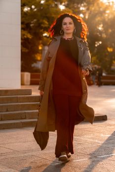 Outdoor fashion portrait of young elegant fashionable brunette woman, model in stylish hat, choker and light raincoat posing at sunset in European city