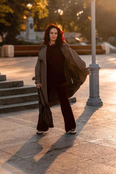 Outdoor fashion portrait of young elegant fashionable brunette woman, model in stylish hat, choker and light raincoat posing at sunset in European city