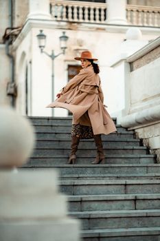 Outdoor fashion portrait of young elegant fashionable brunette woman, model in stylish hat, choker and light raincoat posing at sunset in European city