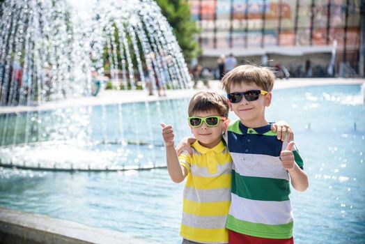 Group of happy children playing outdoors near pool or fountain. Kids embrace show thumb up in park during summer vacation. Dressed in colorful t-shirts and shorts with sunglasses. Summer holiday concept.