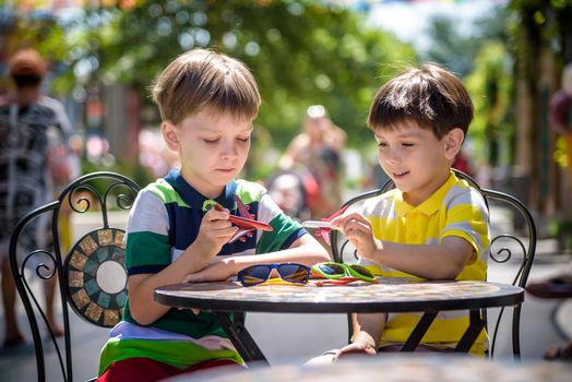 Two little kid boys waiting on table for healthy breakfast in hotel restaurant or city cafe. Child sit on comfortable chair play with toy aircraft, relaxed, enjoy their vacation. Summer holiday with children concept.