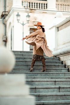 Outdoor fashion portrait of young elegant fashionable brunette woman, model in stylish hat, choker and light raincoat posing at sunset in European city