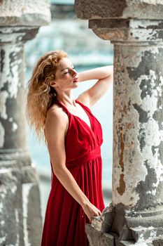 Outdoor portrait of a young beautiful natural redhead girl with freckles, long curly hair, in a red dress, posing against the background of the sea