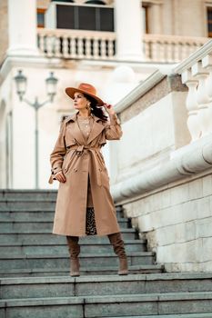 Outdoor fashion portrait of young elegant fashionable brunette woman, model in stylish hat, choker and light raincoat posing at sunset in European city