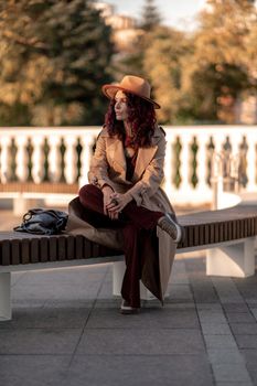 Outdoor fashion portrait of young elegant fashionable brunette woman, model in stylish hat, choker and light raincoat posing at sunset in European city