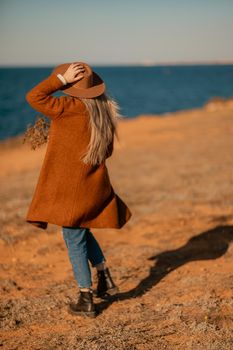 A woman walking along the coast near the sea. An elegant lady in a brown coat and a hat with fashionable makeup walks on the seashore.