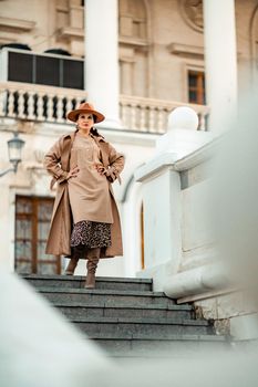 Outdoor fashion portrait of young elegant fashionable brunette woman, model in stylish hat, choker and light raincoat posing at sunset in European city