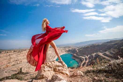 Side view of a beautiful sensual woman in a red long dress posing on a rock high above the lake in the afternoon. Against the background of the blue sky and the lake in the form of a heart.