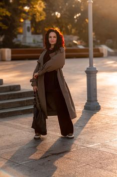 Outdoor fashion portrait of young elegant fashionable brunette woman, model in stylish hat, choker and light raincoat posing at sunset in European city