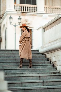 Outdoor fashion portrait of young elegant fashionable brunette woman, model in stylish hat, choker and light raincoat posing at sunset in European city