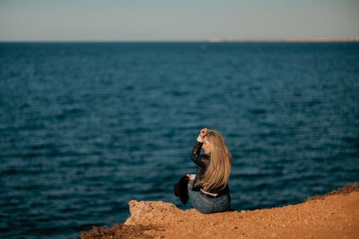 A blonde girl in a stylish black leather jacket is sitting with her back to the seashore